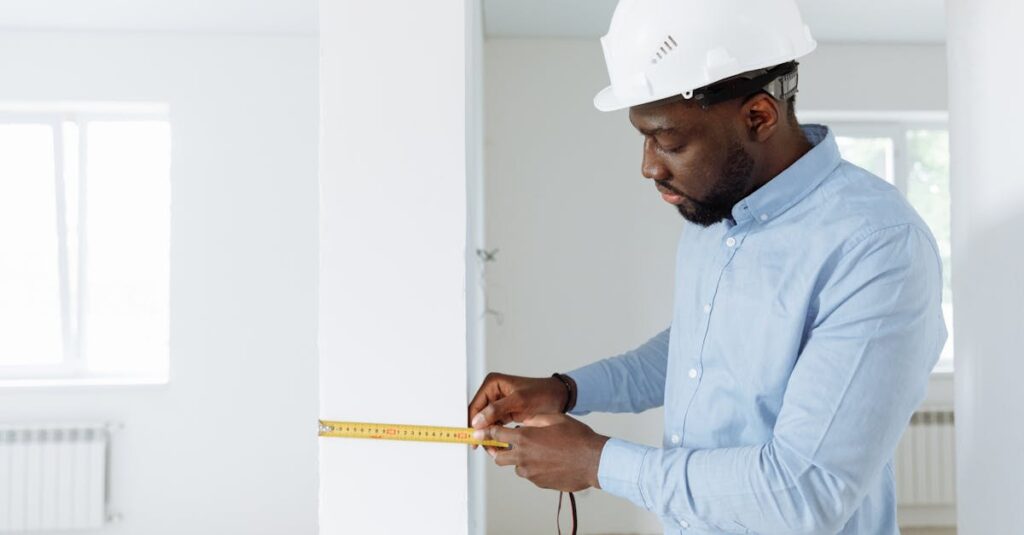 Construction worker measuring a wall indoors, wearing a white hard hat and blue shirt.